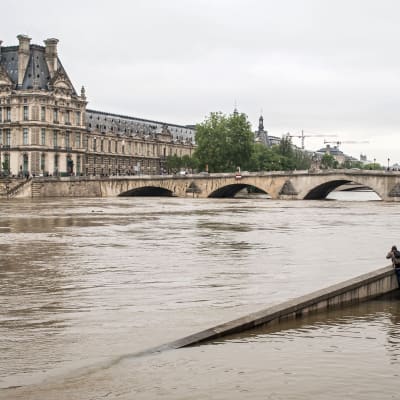 Människor står vid en översvämmad bro och ser över den översvämmade Seine och på museet Louvre.