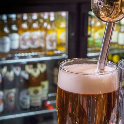 Två ölglas, med öl i ett av dem. I bakgrunden syns ölflaskor i kylskåp.