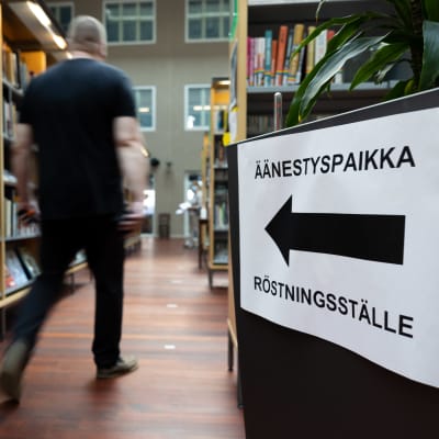 Oikealla kuvassa kyltti, jossa lukee "äänestyspaikka". Mies kävelee käytävää pitkin kyltin takana. 