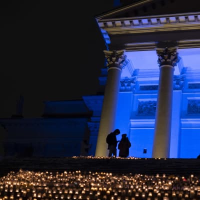 Tuomiokirkon portaille on laskettu paljon kynttilöitä. Rakennusta valaisee sininen valo. Portaiden yläpäässä kahden ihmisen siluetti.