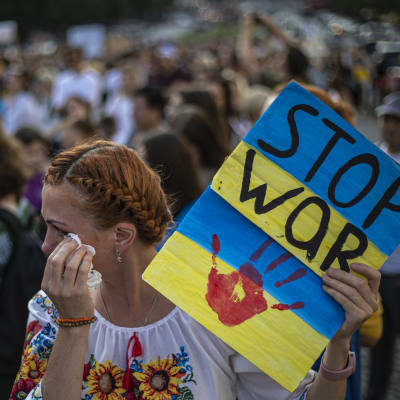 Kvinna med håret uppsatt i en fläta ser bort från kameran och torkar sina tårar med en pappersnäsduk. I handen håller hon en blågul skylt där det står stoppa kriget.
