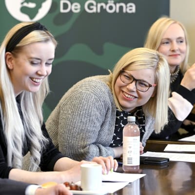 Oras Tynkkynen, Sofia Virta, Saara Hyrkkö och Maria Ohisalo på rad vid ett bord med De Grönas banderoll i bakgrunden. 