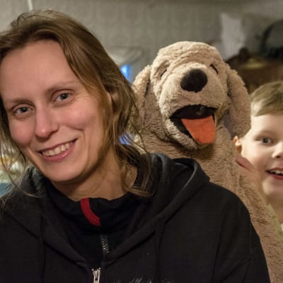 En kvinna ler i förgrunden, bakom henne en pojke med stor leksakshund.