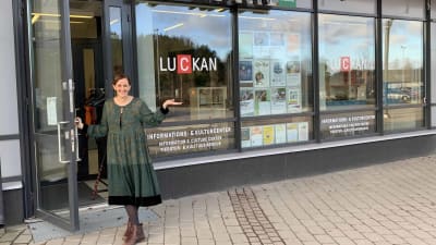 Jenny Paajes står och håller upp glasdörren till Luckan i Kyrkslätt. I fönstret syns Luckans logo, och Jenny är klädd i en grön klänning. Hon ler, och ser in i kameran.