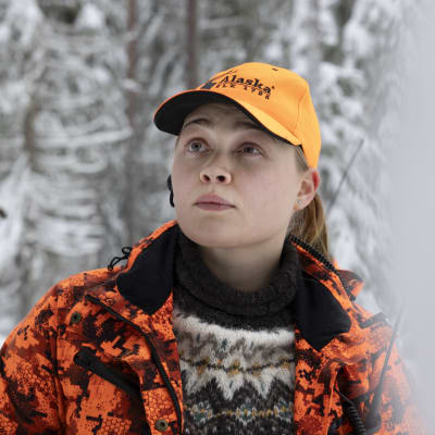 Jutta Jääskeläinen katselee lumisessa metsässä. Hänellä on yllään oranssit metsästysvaatteet ja villapaita. 