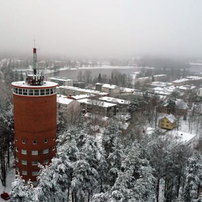 Vanha, punatiilinen vesitorni keskellä lumista mäntymetsää, taustala näkyy Heinolan kaupunki. 