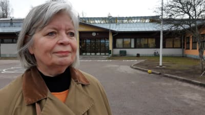 Arja Alho, en äldre kvinna med grått hår och brun rock står på en skolgård.
