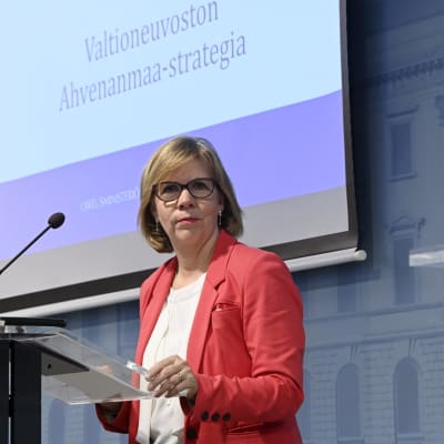 Oikeusministeri Anna-Maja Henriksson seisoo puhujanpöntön edessä, taustalla näkyy näyttö, jossa lukee "Valtioneuvoston Ahvenanmaa-strategia".  
