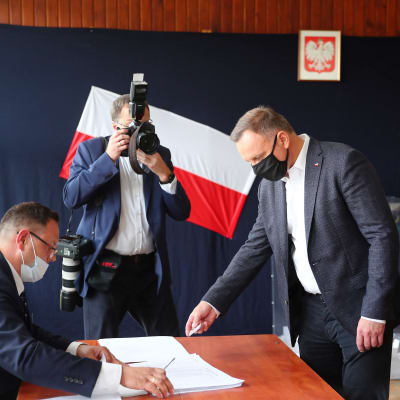 President Andrzej Duda röstar i presidentvalet i Polen den 12 juli 2020. En man ger en valsedel till en annan man, båda bär munskydd. En tredje man fotograferar.