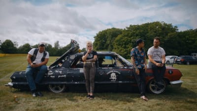 Matha lutar sig mot en svart, amerikansk bil och röker, bredvid henne står och sitter tre män. Bilen är parkerad på en äng, i bakgrunden syns träd och en blå himmel.
