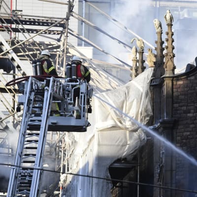 Två brandmän på en lyftkran sprutar vatten över ett rykande hus. 