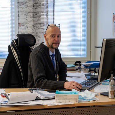 Kannonkosken kunnanjohtaja Sakari Varala istuu työhuoneessaan tietokoneen ääressä ja katsoo hymyillen kameraan silmälasit otsallaan.