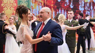 En älde man (Lukasjenko) dansar med en ung kvinna tillsammans med flera andra dansande par i en pråligt dekorerad sal.