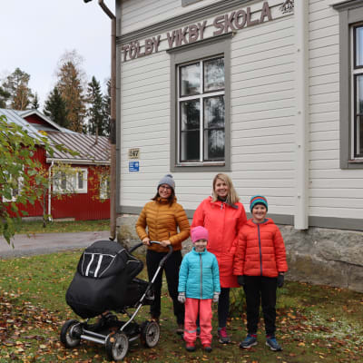 Två kvinnor, ena med barnvagn, och två barn står framför en skolbyggnad.