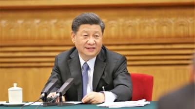 Presidentti Xi Jinping istuu pöydän takana ja puhuu mikrofoniin. Hänellä on edessään papereita. Presidentillä on tummanharmaa puku ja vaalenasininen kravatti.