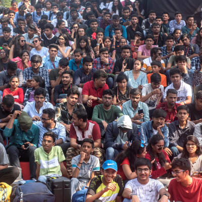 En stor grupp indiska studerande sitter som publik på ett utomhusevenemang.