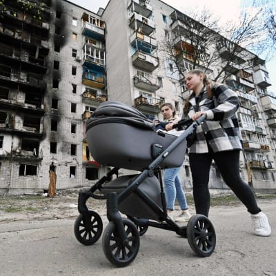 Kaksi nuorta aikuista kävelee kadulla, toinen työntää lastenrattaita. Taustalla palanut kerrostalo.