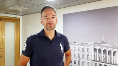 En man i en blå pikétröja. Gråspräckligt skägg och ljusbrunt hår. Står framför en vägg med en tavla och en dörr som har guldfärgad karm.