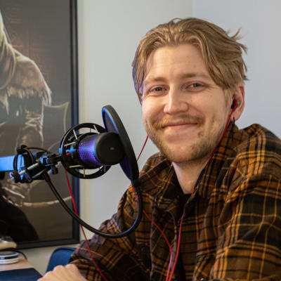 Viking Dufvelin tittar in i kameran och ler. Han står vid sitt skrivbord framför sin mikrofon.