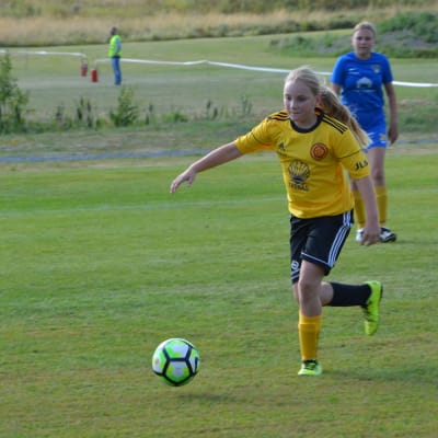 Sonja Karlsson, en fotbollsflicka i gul skjorta sparkar bollen i en matchen. I bakgrunden står lagkamraten Amelie Bärling.