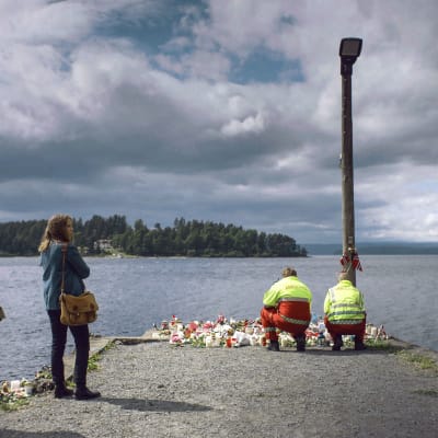 Harald (Marius Lien) och Anine (Alexandra Gjerpen) står vid bryggan mot Utöya, två ambulansmän ställer ner blommor..