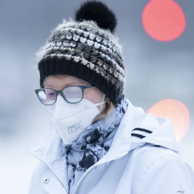 En person i munskydd går ute i vintervädret. Hon har immiga glasögon på sig och mössa.