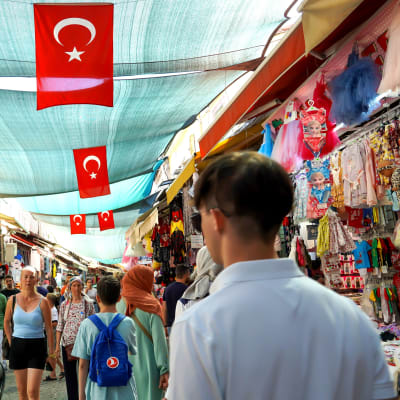 Turkiska flaggor hänger under solskyddstygerna i Kemeralti-basaren i Izmir. Mellan stånden rör sig turister och stadsbor.