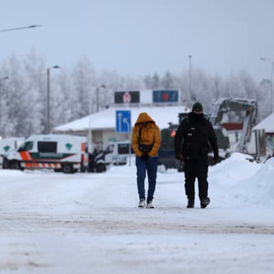 Keltaiseen toppatakkiin ja farkkuihin pukeutunut mies kävelee huppu syvällä päässä, vieressään virkapukuinen henkilö ja taustalla Vartiuksen Venäjän puoleinen rajavartioasema.