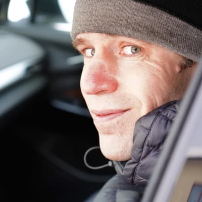 En man med vinterkläder sitter i förarsätet på en personbil och tittar in i kameran.
