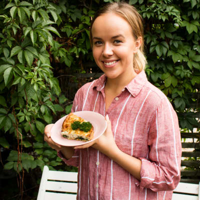 Näyttelijä Sara Parikka seisoo villiviiniseinän edessä ja pitää lautasta kädessään, lautasella on pala piirakkaa. 