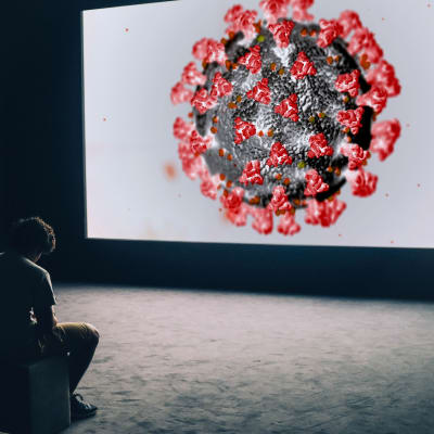 Mies istuu ja tuijottaa skriiniä, jolla näkyy koronavirus.