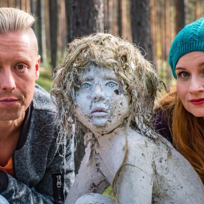 Egenland-programmets programledare Nicke Aldén och Hannamari Hoikkala i skogen tillsammans med en skulptur som ser ut som ett litet barn.