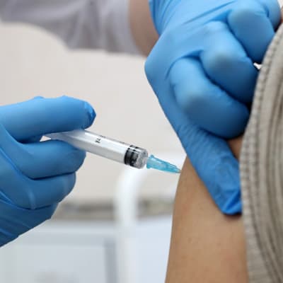 Ett par händer instuckna i blå plasthandskar vaccinerar en person. 