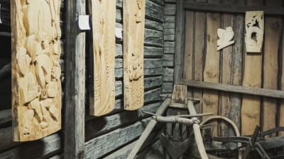 Tre stycken träreliefverk hänger på väggen i en gammal lada. På golvet intill finns gammal släde och seldon som tillhör häst.