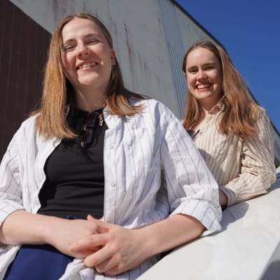 På bilden två unga kvinnor poserar i en trappa i solljus. De står på en trappa och tittar och ler mot kameran.
