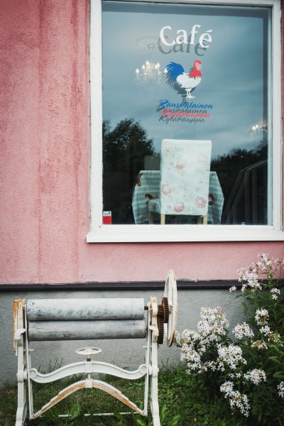 En gammal handvevad mangel står utanför ett chockrosa stenhus. På fönstret texten "Ranskalainen kyläkauppa" och bild på en tupp.