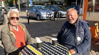 Två leende män sitter på ett torgcafé på Tölö torg.
