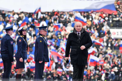 Vladimir Putin kävelee tummassa päällystakissa esiintymislavalla Lužnikin stadionilla.  Hänellä on oikeassa kädessään mikrofoni. Hänestä vasemmalle seisoo kolme univormupukuista ihmistä: kaksi miestä koppalakeissa ja nainen. Taustalla näkyy yleisöä ja Venäjän lippuja katsomossa.