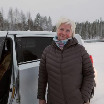 Tumman toppatakkiin pukeutunut nainen, Marina Määttä, seisoo autonsa vieressä, taustalla luminen havumetsä Vartiuksessa.
