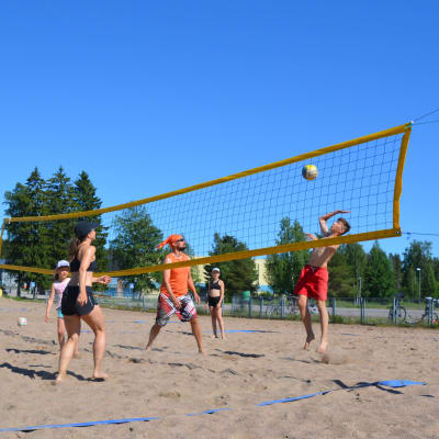 En grupp människor i olika åldrar spelar beachvolley. En pojke hoppar upp vid nätet och ska slå bollen.