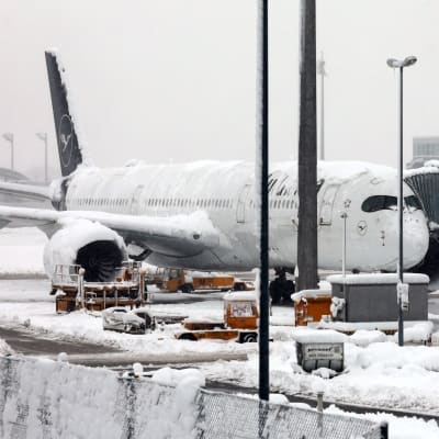 Ett Lufthansaplan står vid en gate med snö på flygplanskroppen och vingarna. Omkring planet är det också snö på mark och fordon.