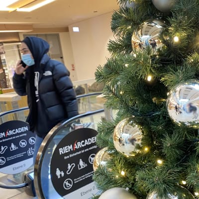 Bilden är tagen i ett köpcenter. Närbild på en julgran med lampor och silvriga stora julgransbollar. I bakgrunden ser man en rulltrappa som andänds av en person med uppdragen huva och munskydd.