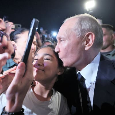 Putin suukottaa nuorta naista väkijoukossa. Ihmiset ottavat kuvia kännykkäkameroilla.