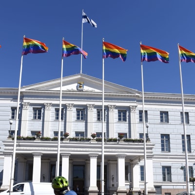 En person cyklar förbi stadshuset i Helsingfors. På taket fladdrar regnbågsflaggor.
