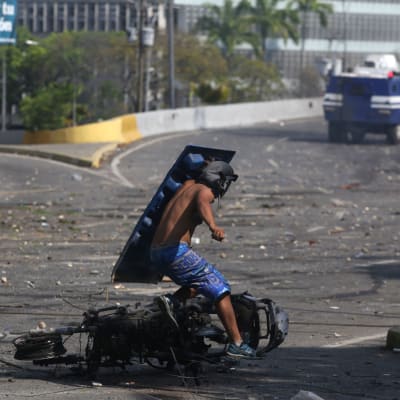 En sköldförsedd demonstrant springer förbi en utbränd motorcykel under kravellerna i Caracas 