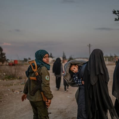 Två hustrur till IS-krigare går längs en väg. Bredvid dem går en kvinnlig kurdisk soldat.