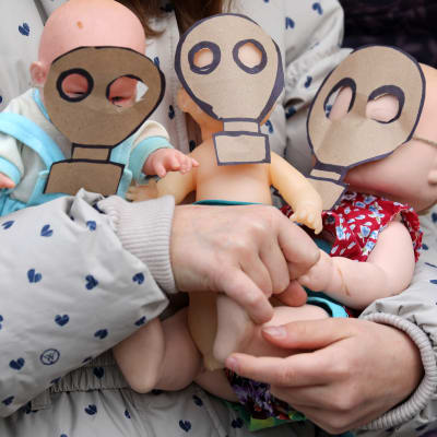 Lapsi pitelee sylissään kolmea vauvanukkea, joiden kasvoilla on paperiset kaasunaamarit.