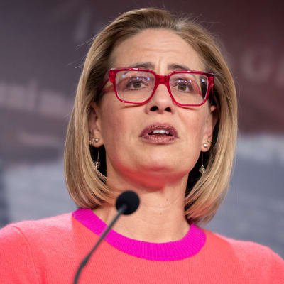 En blond kvinna med röda glasögon talar med mikrofoner framför sig. Hon ser orolig ut.