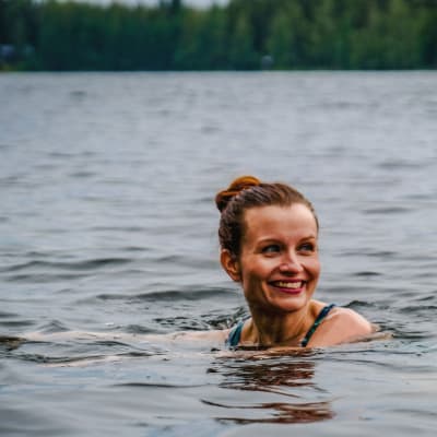 Nainen uimassa järvessä, näkyy vain pää, katsoo hymyillen ohi kameran