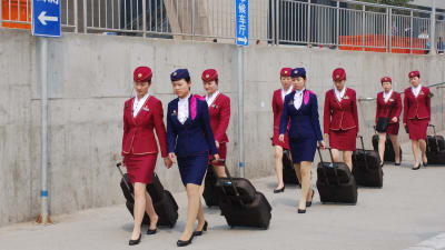 Kinesiska tågvärdinnor i sina blåa och röda kostymer på väg mot jobbet. I uniformen ingår högklackade svarta skor och en liten hatt. Värdinnorna drar på kappsäckar.
